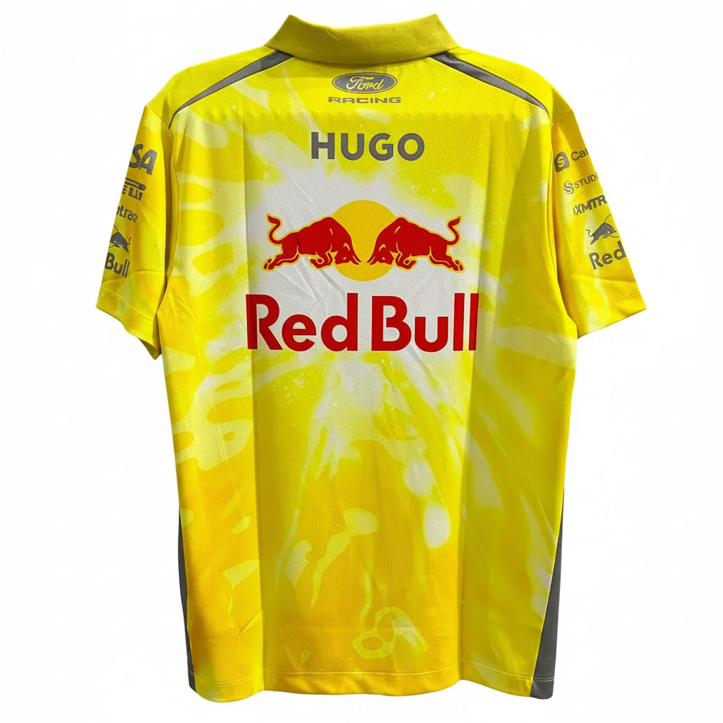 A yellow short-sleeve racing jersey with a white tie-dye pattern, featuring the Red Bull logo and 'HUGO' printed on the back. The jersey is displayed against a decorative white wall with black leaf-like cutouts.