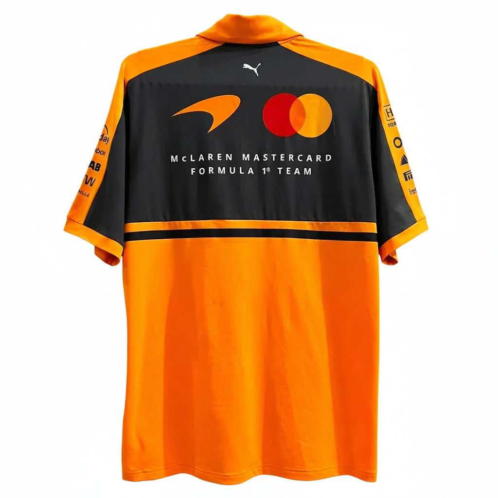 A short-sleeve McLaren M1astar Formula 1 Team Puma jersey shown from the back, featuring a black upper back with sponsor logos and a bright orange lower body and collar, hanging against a decorative white screen.