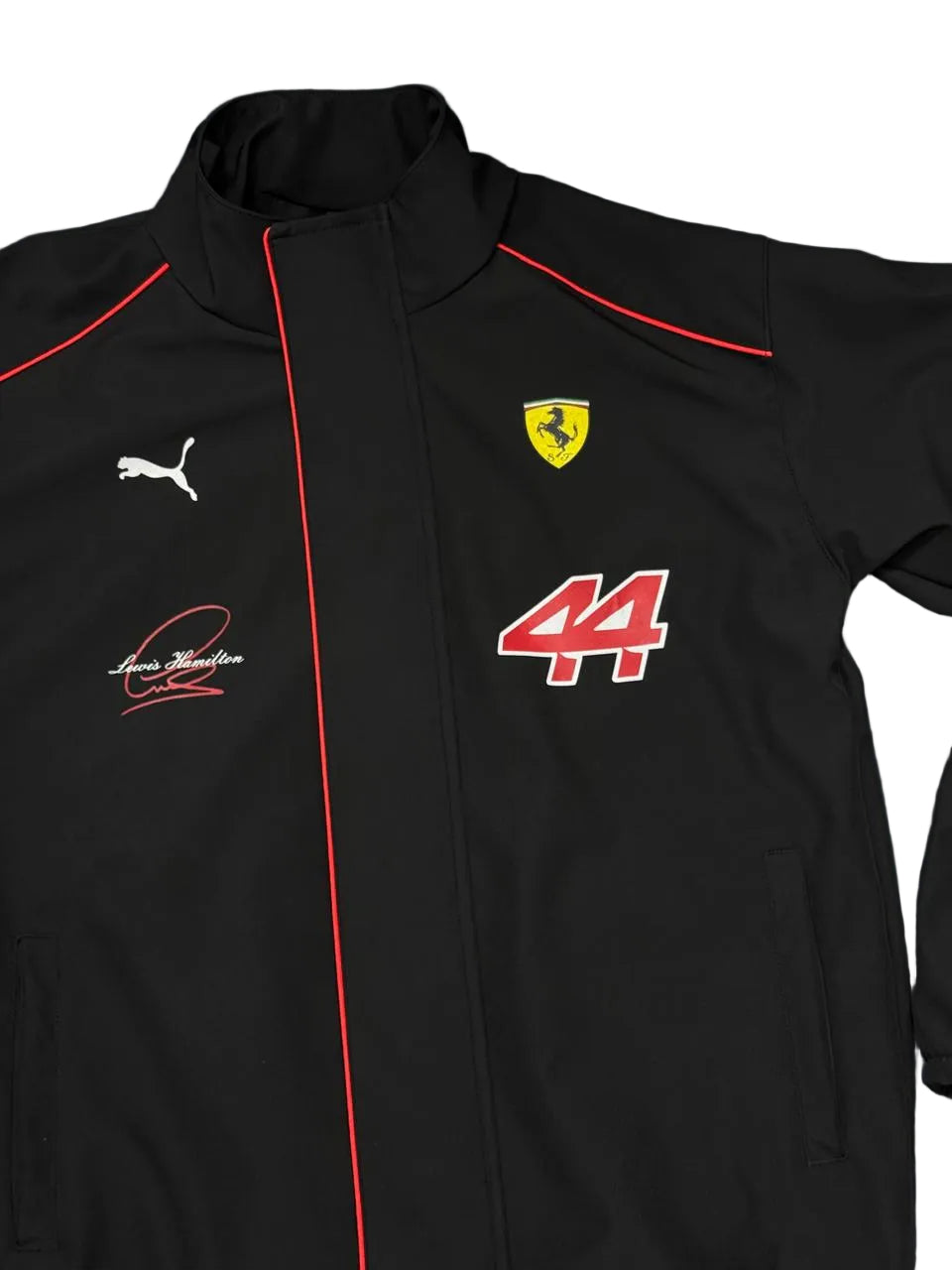 A black Puma Ferrari zip-up jacket featuring red piping, the yellow Ferrari shield logo, a white Puma cat logo, and a red and white number 44 on the chest, laid flat on a dark blue fabric background.