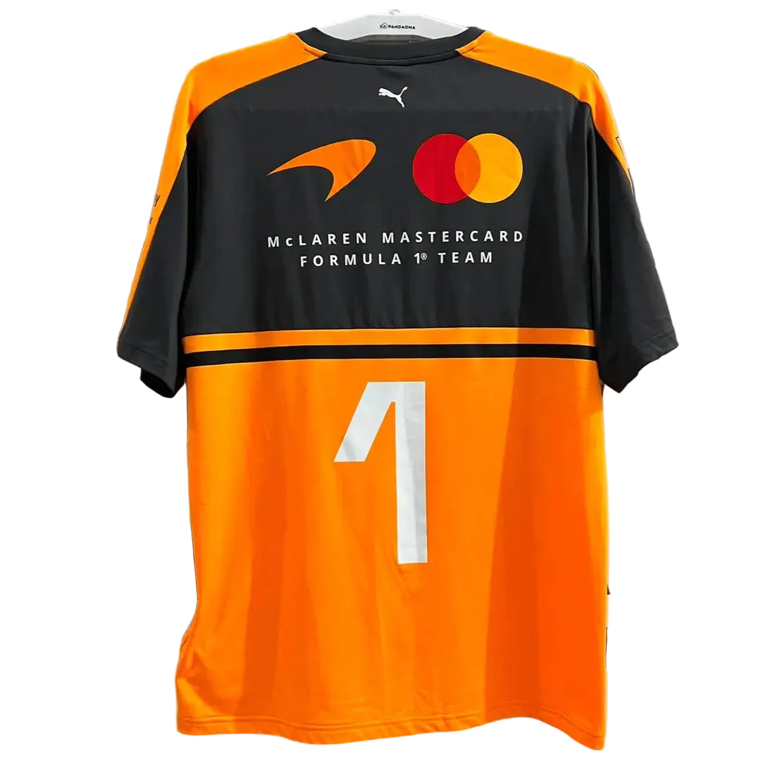 A black and orange McLaren Mastercard Formula 1 Team jersey with the number 1 on the back, featuring the Puma logo and sponsor logos, hanging against a decorative white screen.