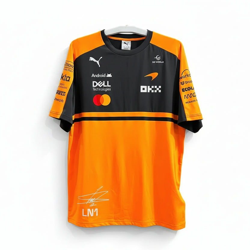 A short-sleeve orange and black Puma McLaren Mercedes F1 team shirt with sponsor logos, featuring a signature print on the lower front, hanging on a white hanger against a decorative black-and-white patterned wall.