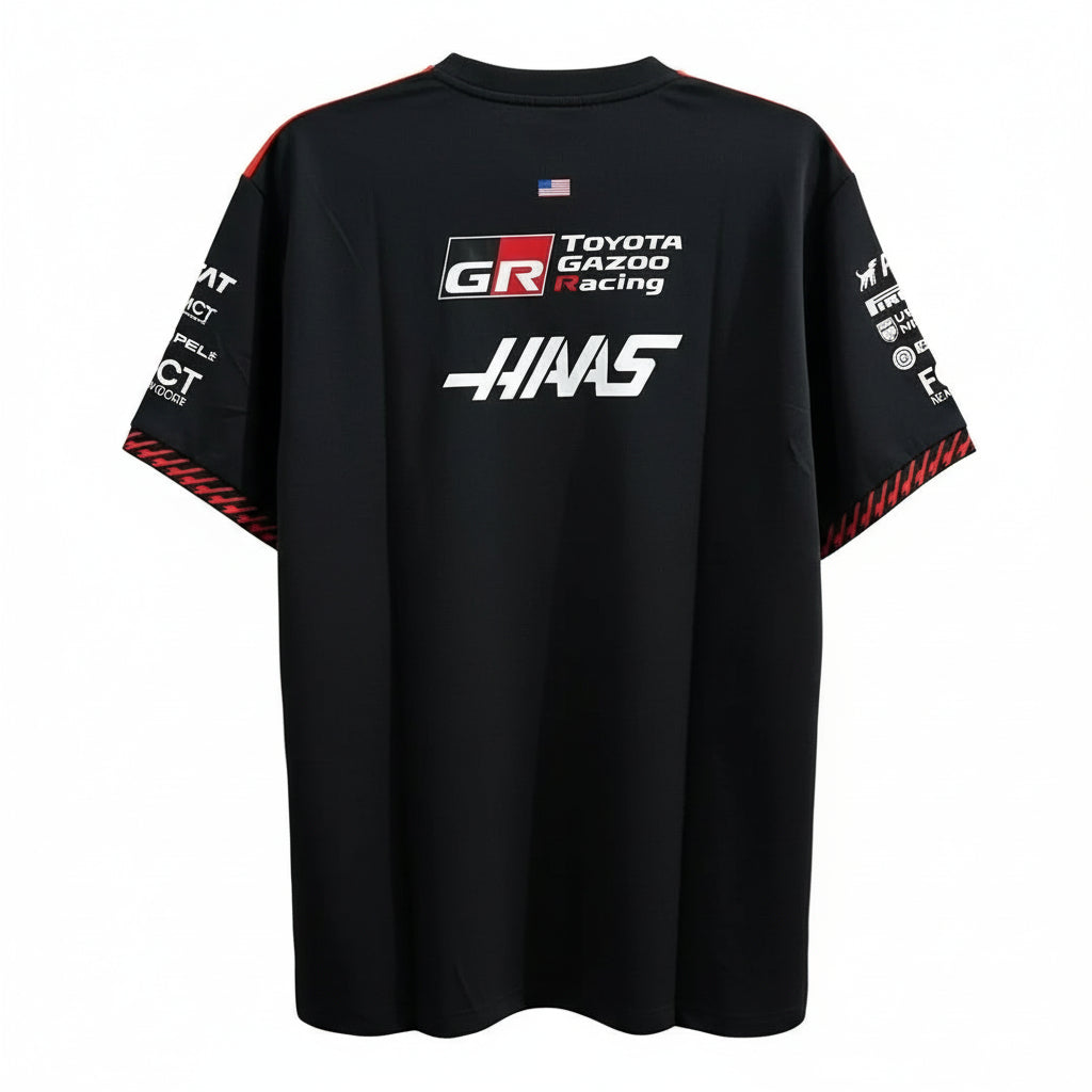 A black short-sleeve T-shirt with red and white graphics, featuring 'Toyota Gazoo Racing' and 'Haas' logos on the back, hanging against a white decorative screen.
